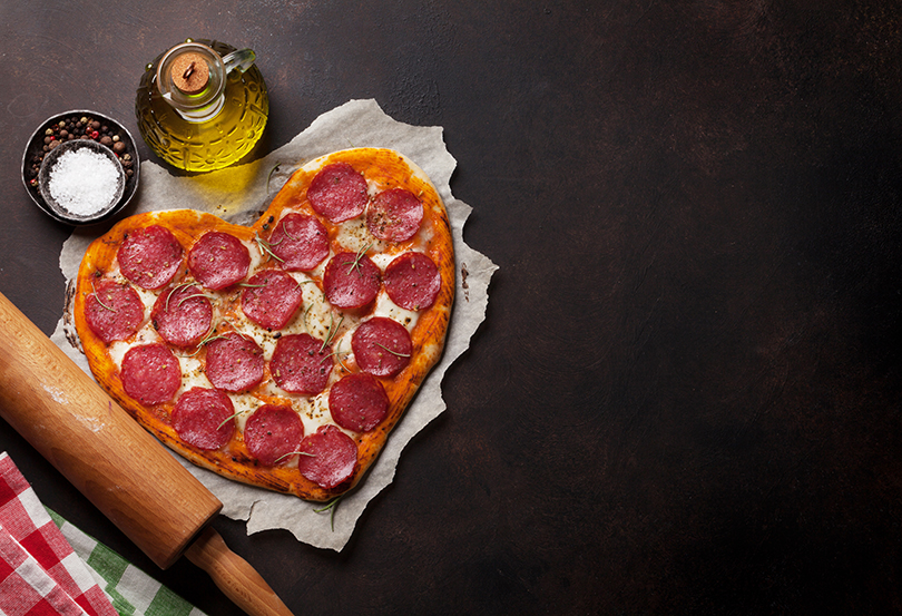 Pizza de calabresa em formato de coração, em uma bancada escura com elementos de culinária incorporando a imagem.