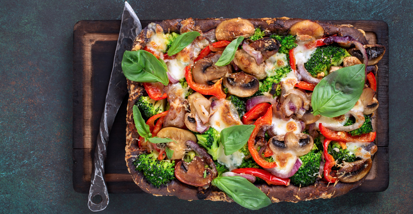 Pizza retangular com recheio de legumes coloridos, sobre uma tábua de madeira escura e uma faca de inox.
