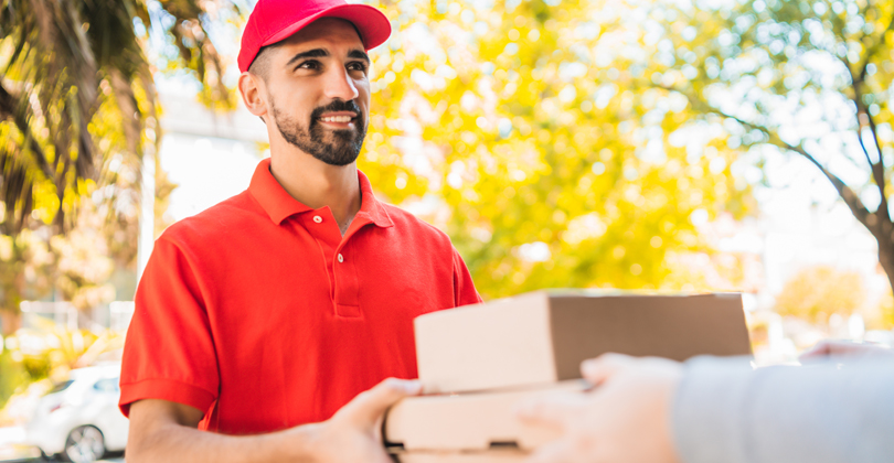 Homem realizando uma entrega de pedido feito via delivery, vestindo camiseta e boné vermelhos, sorrindo.