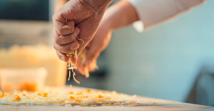 Mãos de um pizzaiolo colocando queijo sobre uma pizza de milho.