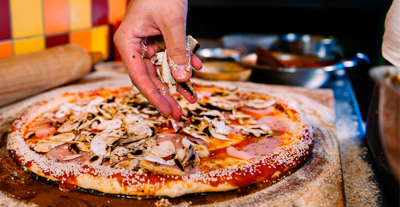 Mãos de um pizzaiolo distribuindo cogumelos sobre uma pizza com borda de gergelim